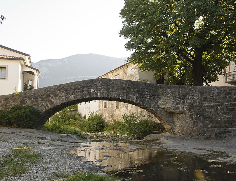 Old stone arch bridge spanning a small, clear stream in a traditional village, showcasing historic architecture and the serene countryside of Slovenia tourism.