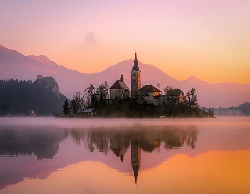 Iconic sunrise over Bled Island and its church with mist covering the lake and pink reflections; a key destination for tourism in Slovenia.