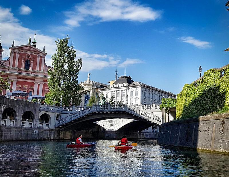 Two kayakers paddling on the Ljubljanica River under the Cobblers' Bridge in Ljubljana, with a view of the Franciscan Church; promoting urban activities in Slovenia tourism.