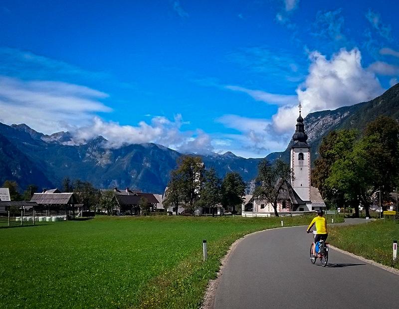 Cyclist on a self-guided bike tour riding past St. John the Baptist Church in Bohinj with mountains in the background, combining culture and nature in Slovenia.