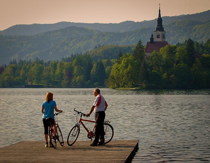 Couple on a Helia self-guided cycling tour standing with bikes on a jetty at Lake Bled, with the iconic island church promoting active Slovenia tourism.
