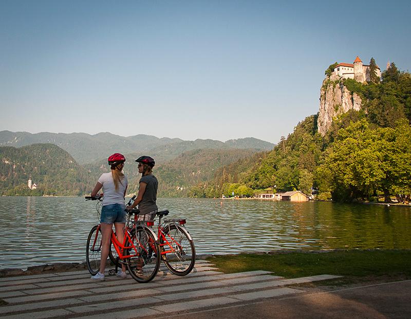 Two female cyclists standing with their rental bikes, looking across Lake Bled toward the historic Bled Castle perched dramatically on the cliff face. Scenic rest spot for self-guided tours.