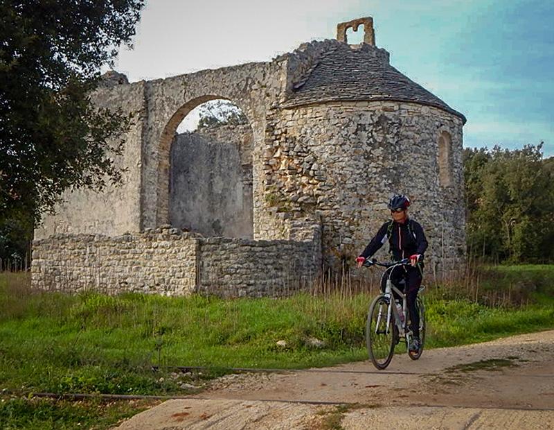 A cyclist on a dirt path next to the historic stone ruins of an ancient chapel or church in the Slovenian Karst region, highlighting cultural stops on a self-guided tour.