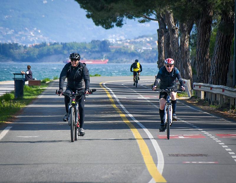 Group of cyclists riding along the scenic coastal road near Koper, Slovenia, with the Adriatic Sea and a port in the background. Perfect for a Helia self-guided coastal tour.