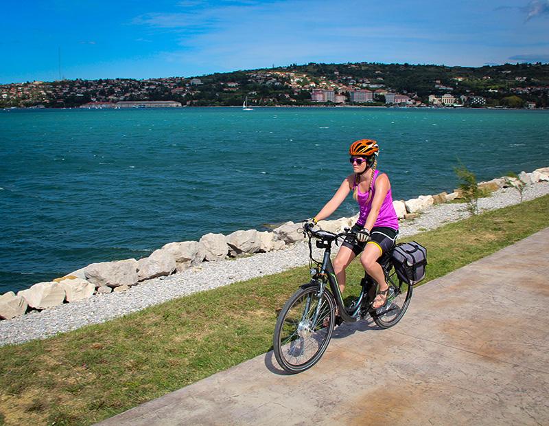 A female cyclist on an e-bike riding a paved path right next to the Adriatic Sea on the Slovenian Coast, with a sunny hillside town in the distance. Self-guided coastal cycling.