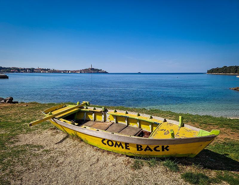 A bright yellow rowing boat with "COME BACK" painted on it sits on the beach, overlooking the historic coastal town of Rovinj, Istria, across the clear Adriatic Sea. A memorable stop on a Helia Istria cycling tour