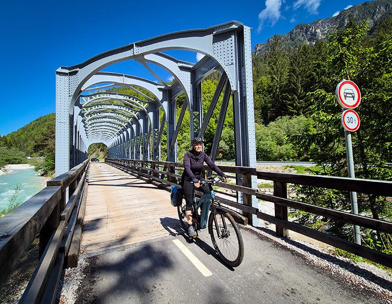 A female cyclist riding an e-bike across a distinctive grey steel arch bridge over the turquoise Sava River in the Julian Alps, Slovenia. A memorable sight on a Helia self-guided tour