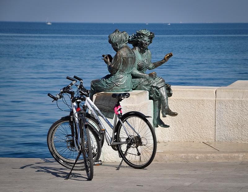 Two touring bikes parked by the famous 'Le Ragazze di Trieste' statue on the waterfront, marking the end of a Helia self-guided cycling tour to the Adriatic Sea