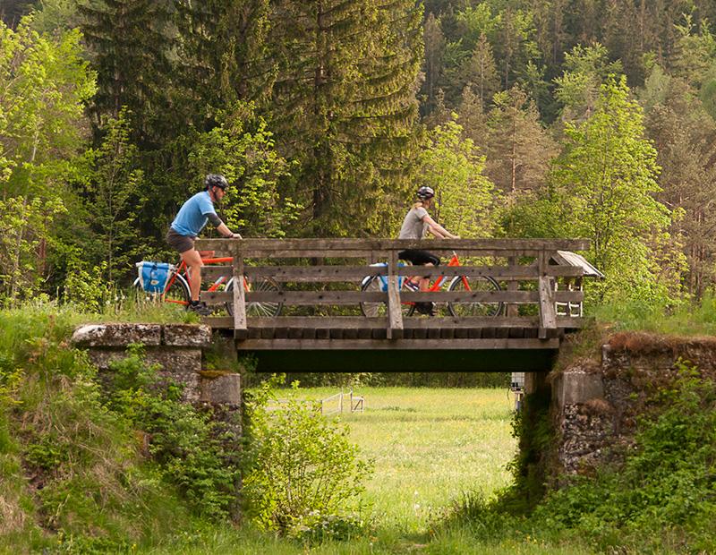 Two cyclists crossing a rustic wooden bridge on a self-guided bike tour through the lush Slovenian countryside with Helia.