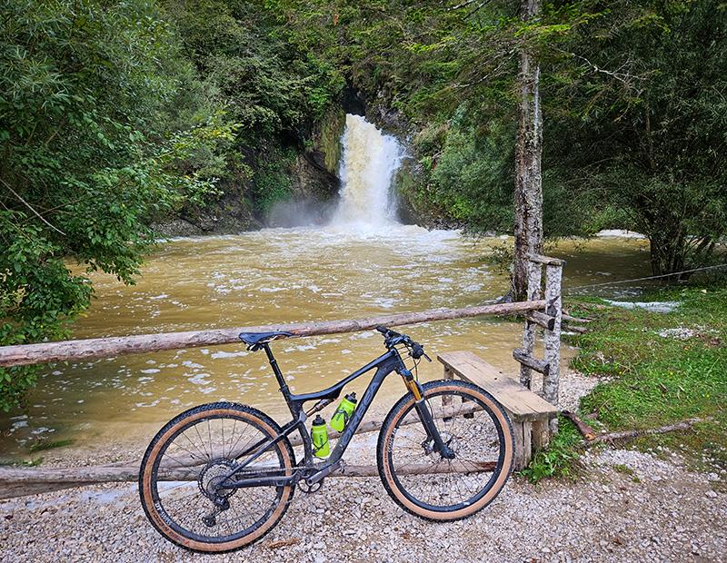 A full-suspension mountain bike parked next to a wooden fence with a powerful, arching waterfall in the background. MTB adventure in Slovenia with Helia.