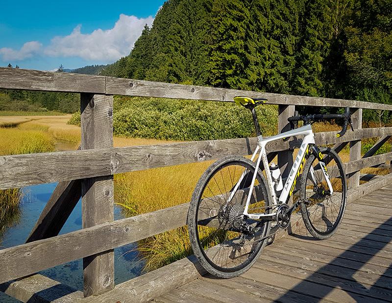 A white and yellow gravel bike resting on a wooden bridge crossing a small stream in a sunny, marshy meadow in Slovenia. Helia adventure cycling.