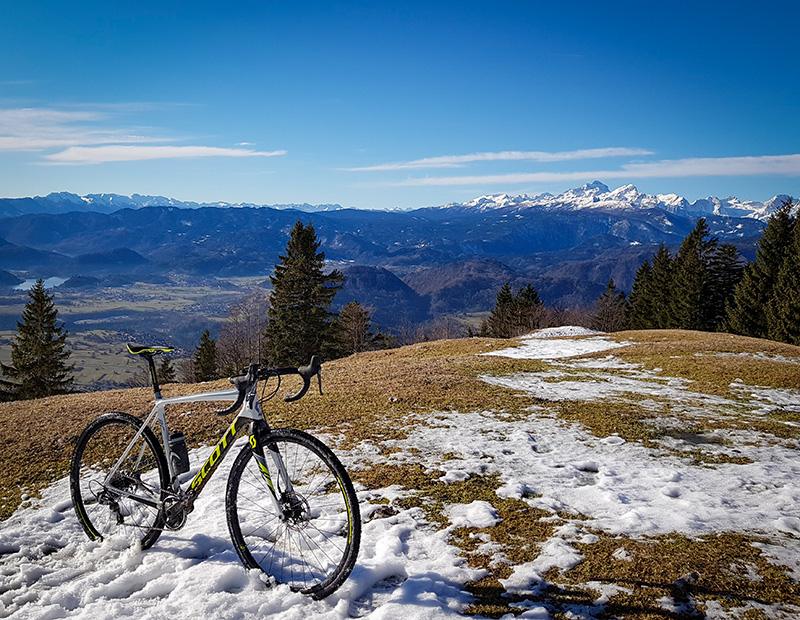 A gravel bike resting on a patch of snow overlooking a valley and the stunning snow-capped Julian Alps mountain range and Bled Valley. Gravel cycling Slovenia