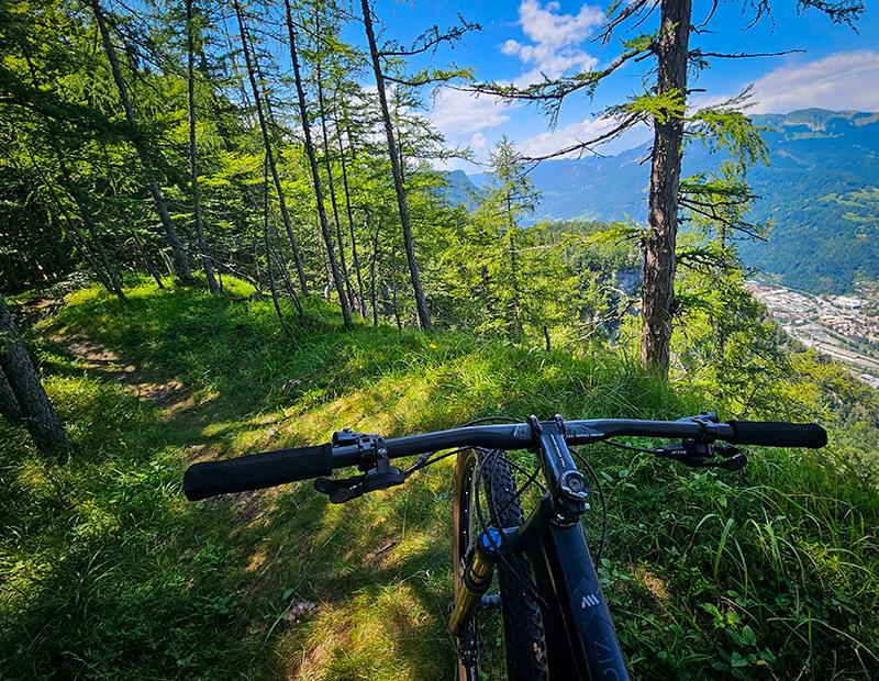 First-person perspective of mountain biking on a singletrack trail high above a valley and town, showing the view from the handlebars. MTB in Slovenia