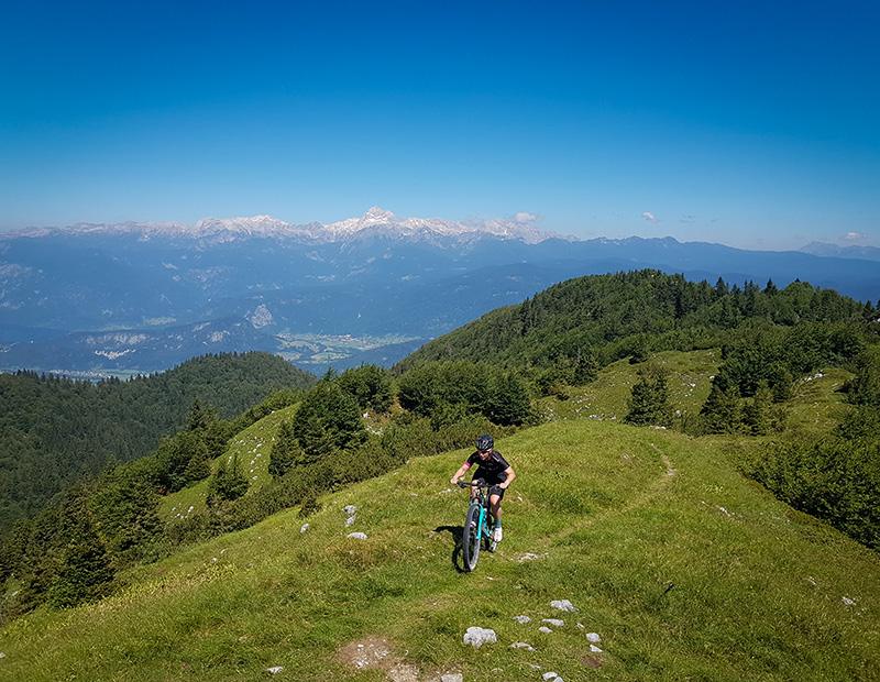 Mountain biker riding a grassy alpine ridge with a spectacular panoramic view of the snow-capped Julian Alps and Mount Triglav, Slovenia. Helia mountain biking tour