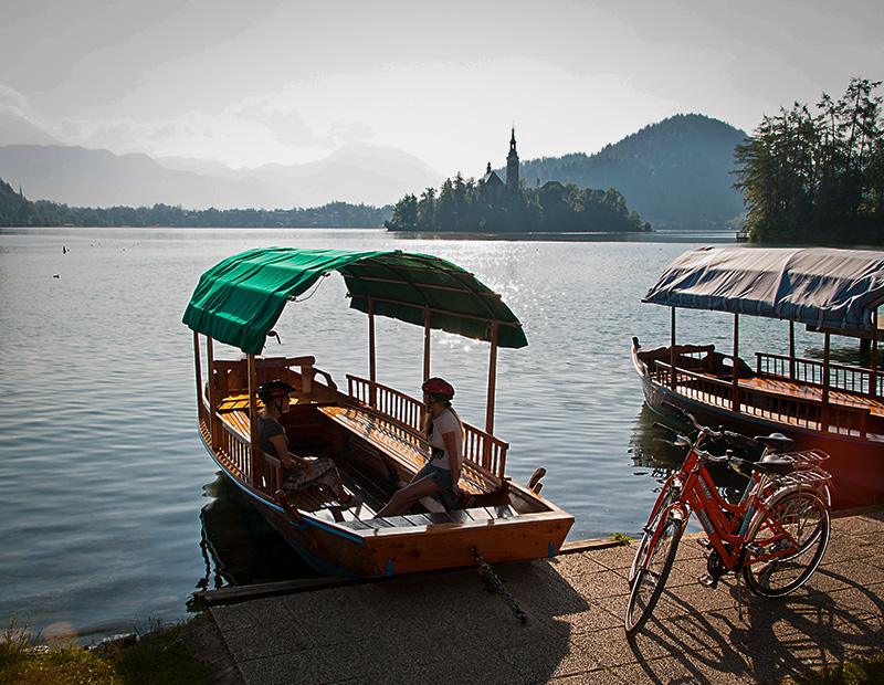 Two cyclists ready to board a traditional Pletna boat on Lake Bled, with an e-bike parked nearby, highlighting cultural stops on a multi-activity cycling tour in Slovenia.