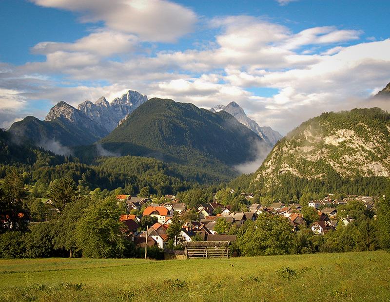 Picturesque alpine village nestled in a green valley, framed by dramatic, misty Julian Alps mountains in Gorenjska, Slovenia. A scenic stop on a Helia cycling holiday.