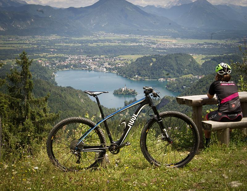 A mountain biker resting on a bench with their bike, overlooking the iconic Lake Bled, the island church, and the surrounding valley. Scenic break on a Helia MTB tour in Slovenia.