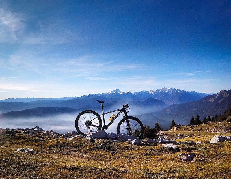A mountain bike resting on a rocky alpine ridge, overlooking a hazy, beautiful layered panorama of the Julian Alps and distant mountains in Slovenia. High-altitude cycling
