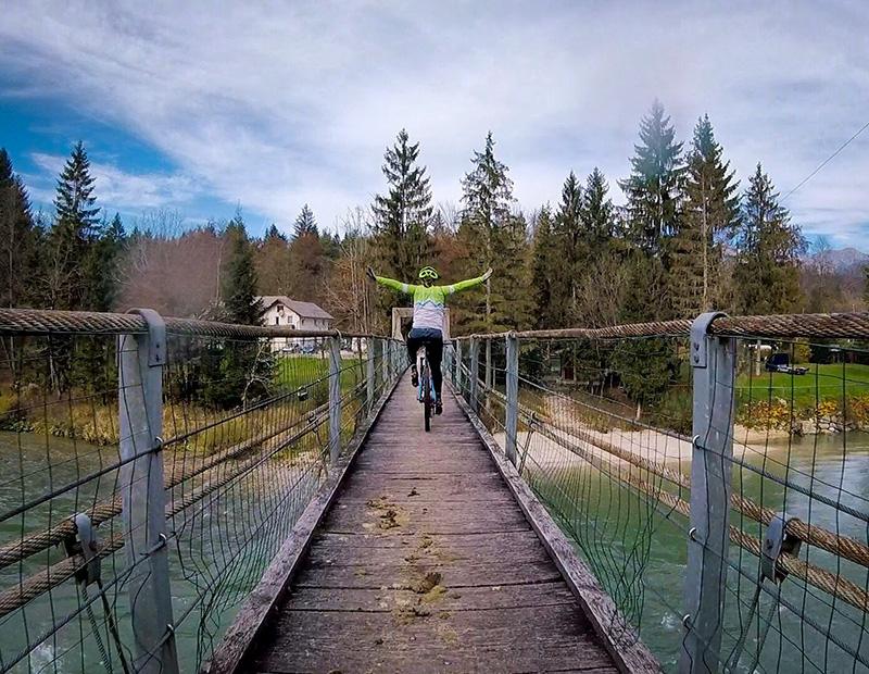 Adventurous cyclist with arms raised riding a wooden suspension bridge over a turquoise river in Slovenia, celebrating the freedom of a Helia adventure cycling tour.