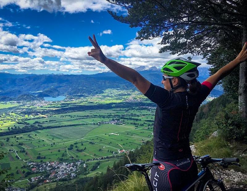 Female mountain biker celebrating with arms raised at a high mountain viewpoint, looking down on a vast green valley and the distant Lake Bled in Slovenia. Helia tour triumph