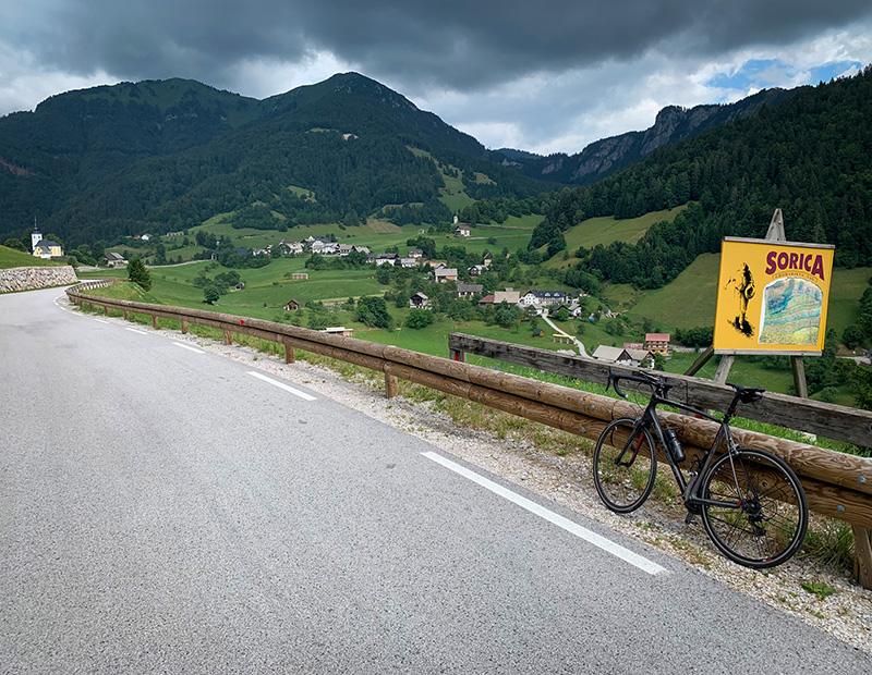 Road bike leaning against a guardrail above the alpine village of Sorica, surrounded by green hills and dramatic mountain scenery in Slovenia