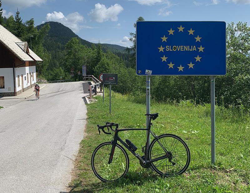 Road bike leaning against a Slovenia border sign on a quiet alpine road, with a cyclist approaching from the distance
