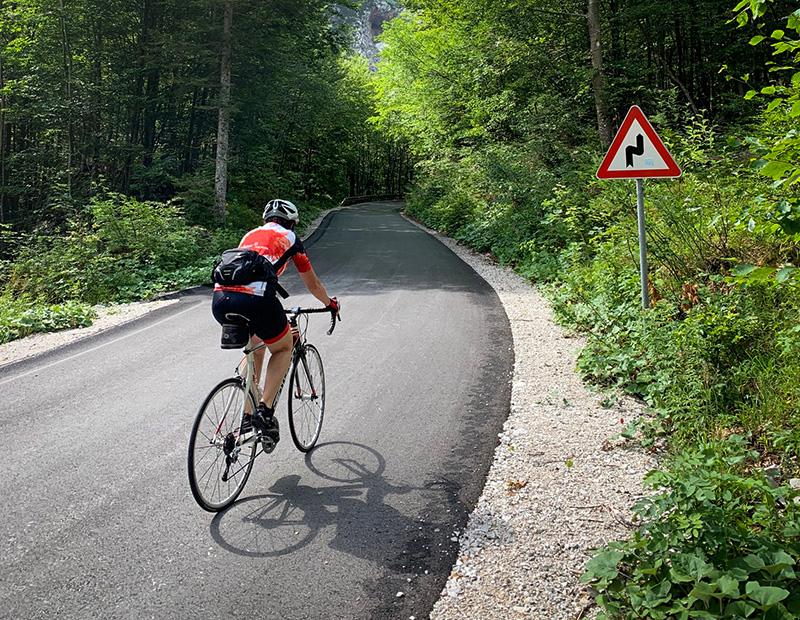 Cyclist riding along a winding forest road on a sunny day, approaching a sharp turns sign in the Slovenian Alps