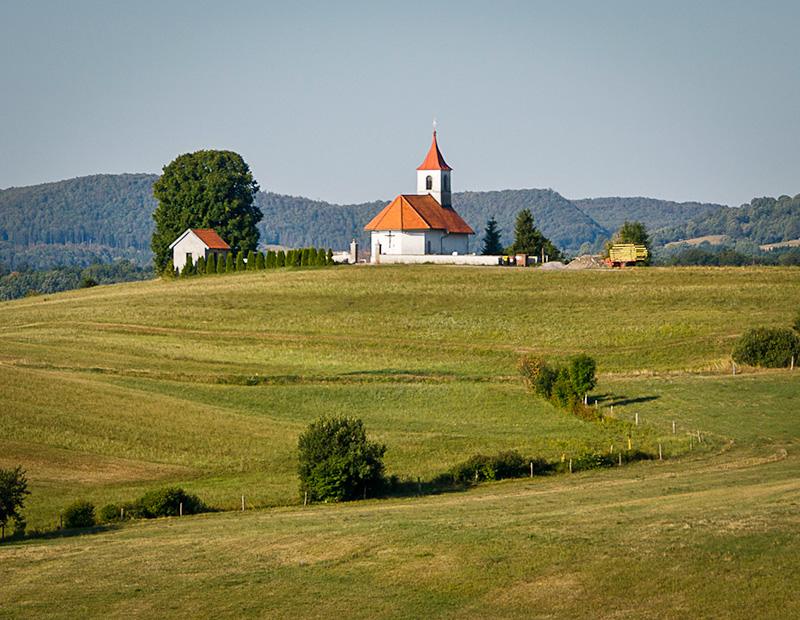 Picturesque small white church with a red roof on a green hill, representing rural cultural heritage and scenic routes for cycling tours in Slovenia tourism.