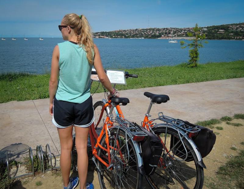 Woman taking a break from cycling, standing with her touring bike, looking out at the sea and coastal view in Slovenia tourism.